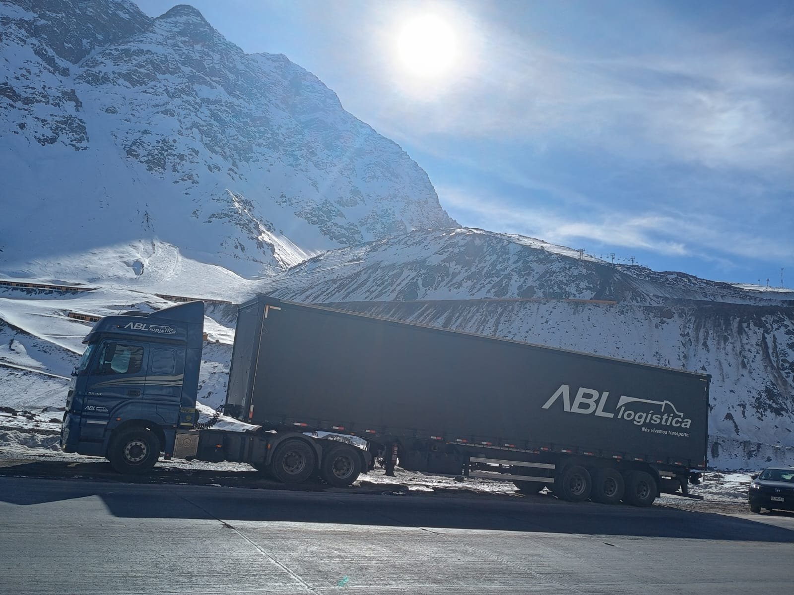 Caminhão azul com carreta cinza da ABL Logística parado em uma estrada asfaltada. Ao fundo, uma imensa montanha coberta de neve sob um céu azul e sol forte.